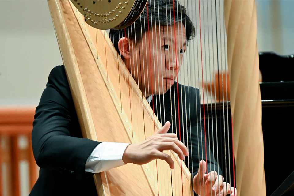A male student, wearing a smart suit, looking at the conductor, while playing the harp.